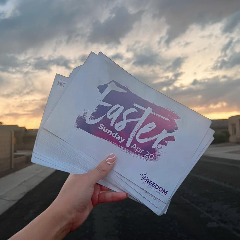 A hand holding Easter invitation cards for Freedom Baptist Church in Yuma, Arizona, against a colorful sunset sky.
