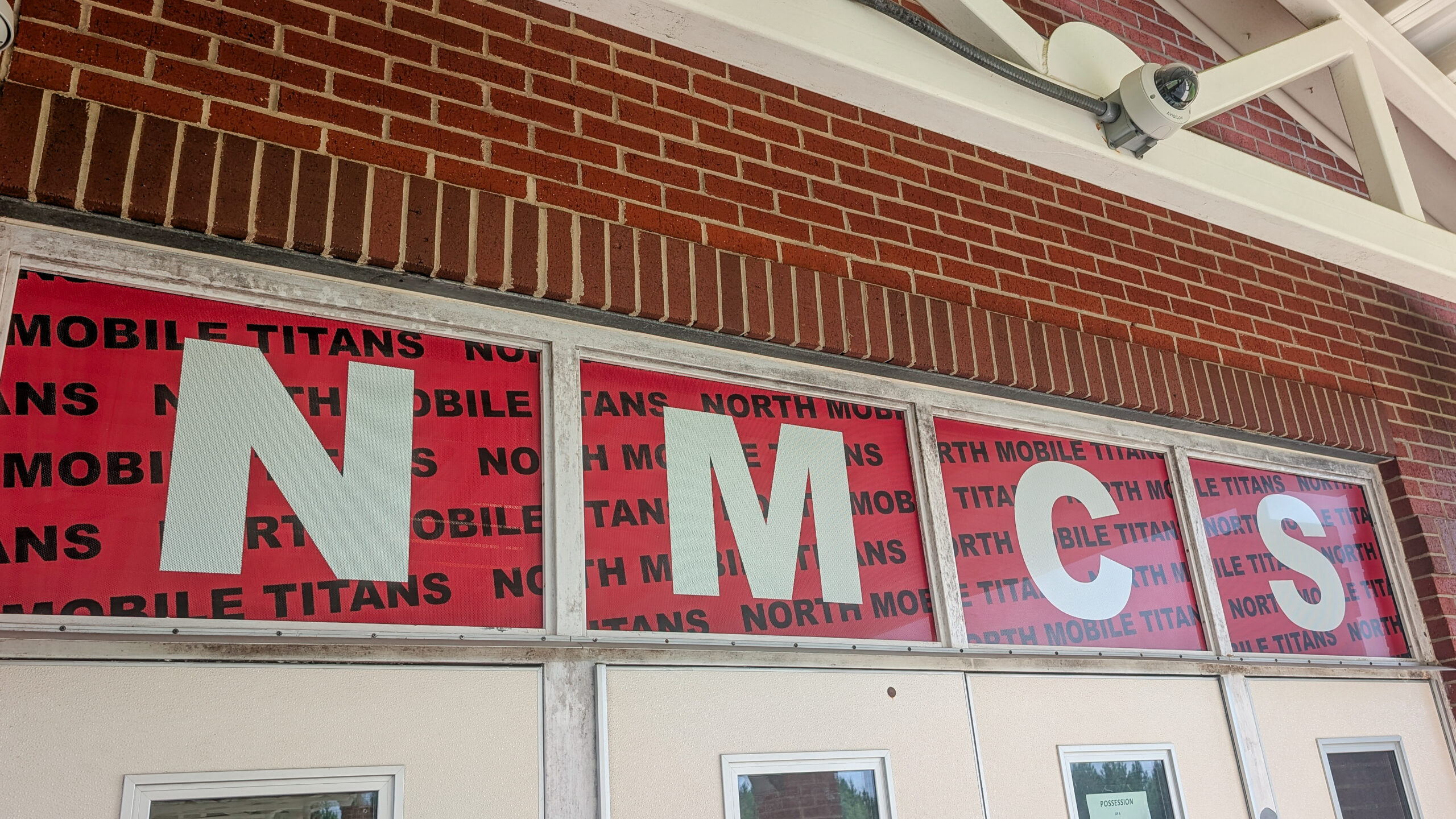 Custom window perforated graphics spelling “NMCS” above the front entrance doors at North Mobile County School in Mobile, Alabama, featuring bold white letters over a red Titan-themed background.